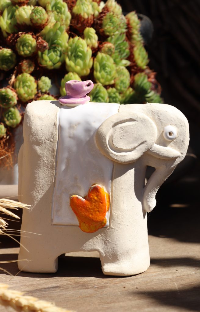 weißer Elefant mit einer rosanen Tasse am Rücken und einem weißen Tuch mit einem orangenen Herz